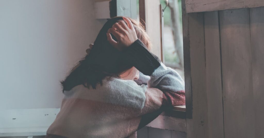 A woman sits indoors gazing thoughtfully out of a window, evoking emotions of loneliness and reflection.