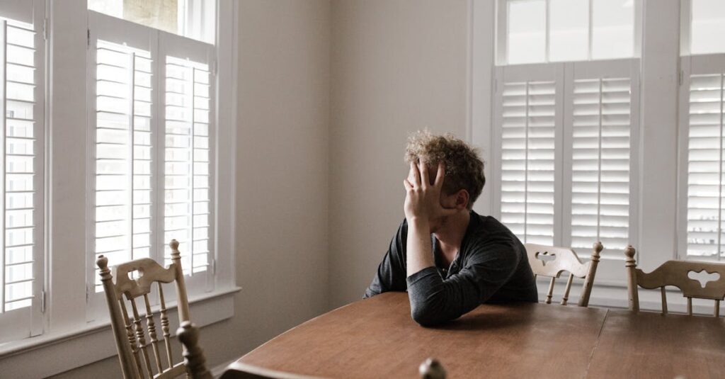 A man sits alone at a table in a bright room, displaying deep contemplation.
