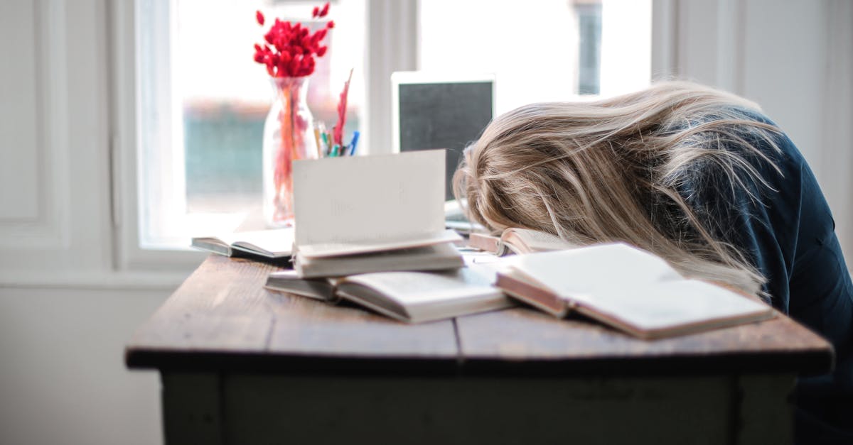 Blonde woman slumped over desk filled with books, showcasing study fatigue indoors.